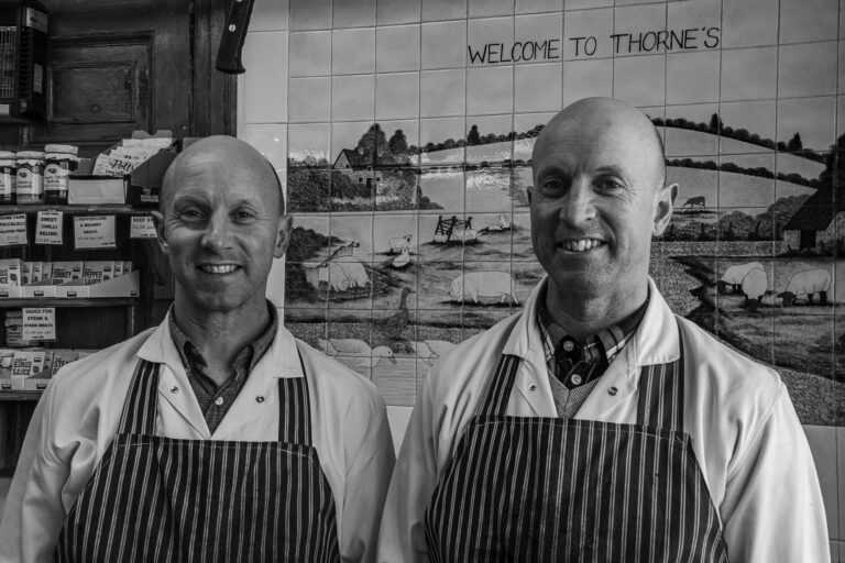 Picture of the twin brothers who run the butcher's shop, both smiling and in their stripy aprons, in front of a tiled shop sign